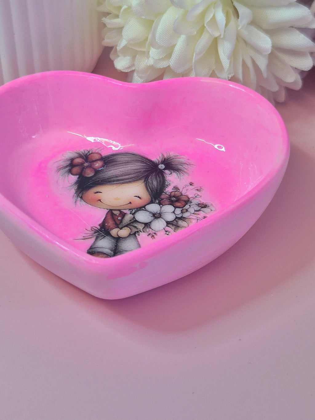 Heart-shaped pink dish with a cartoon character holding flowers on a pink surface with white flowers in the background.