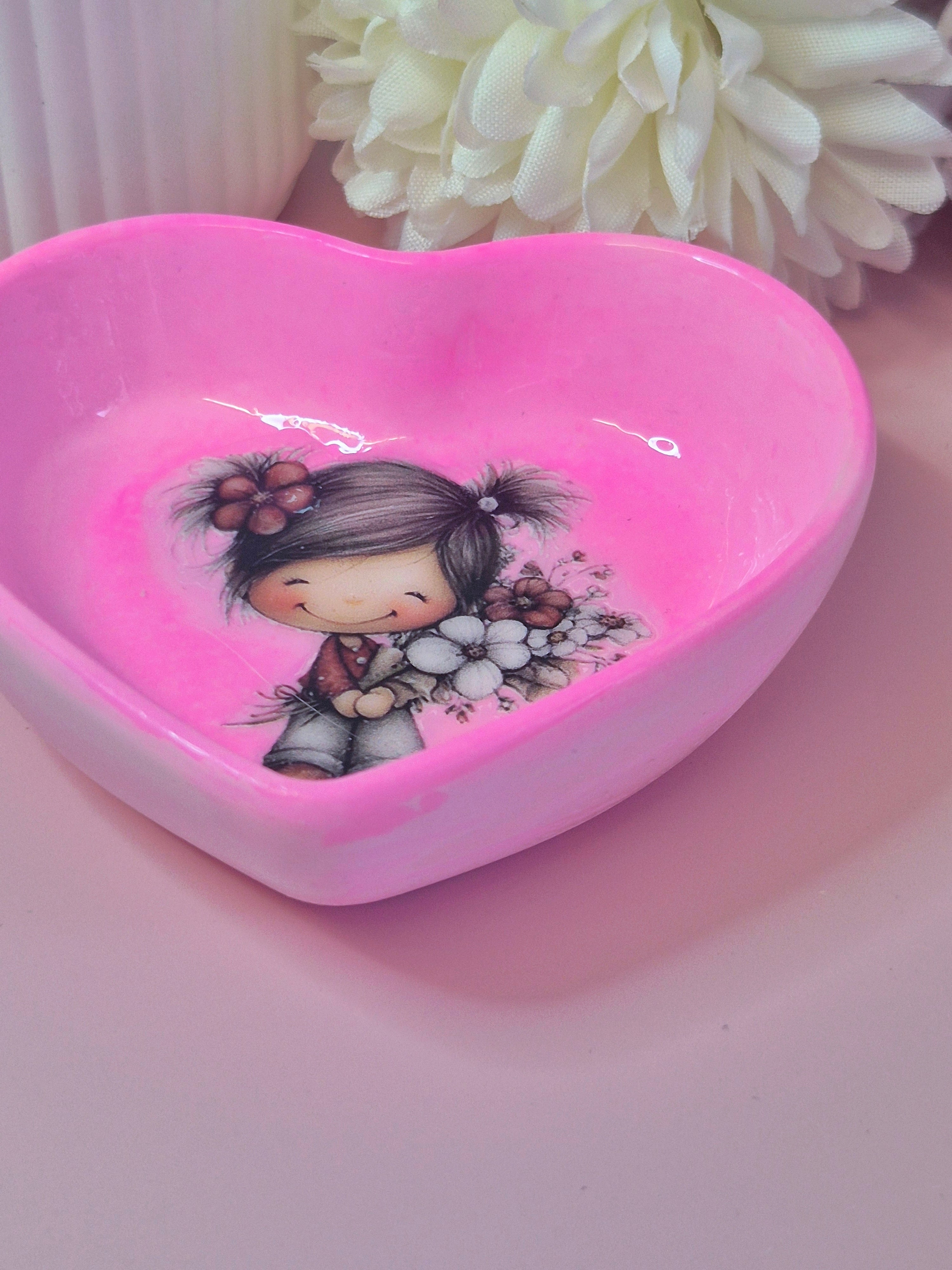 Heart-shaped pink dish with a cartoon character holding flowers on a pink surface with white flowers in the background.