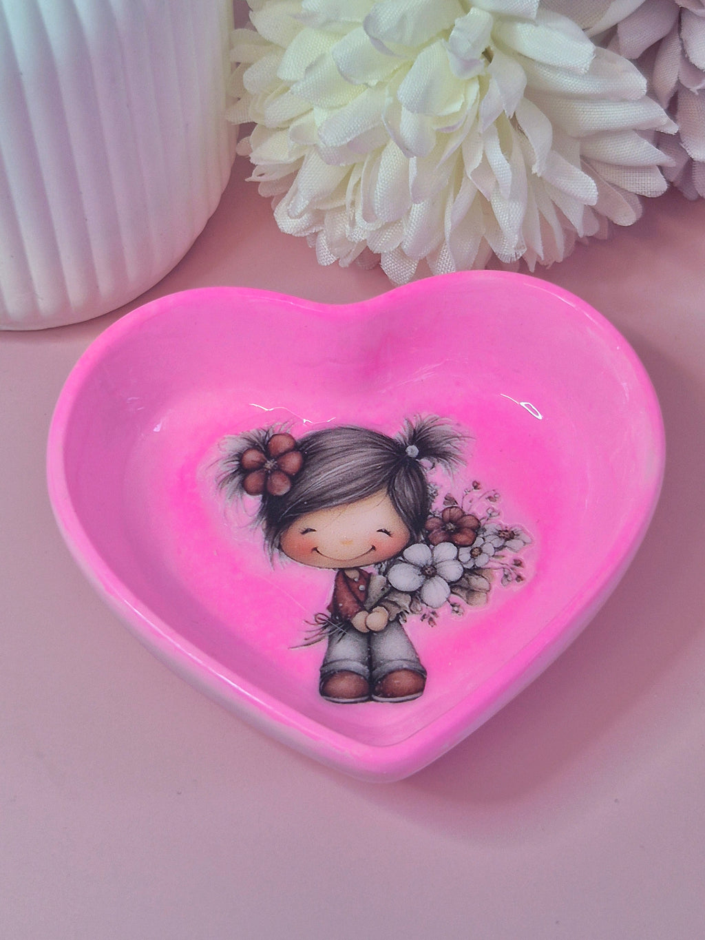 Heart-shaped pink trinket dish with a girl holding flowers on a pink surface with white flowers in the background.