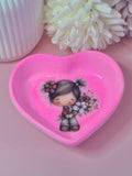 Heart-shaped pink trinket dish with a girl holding flowers on a pink surface with white flowers in the background.