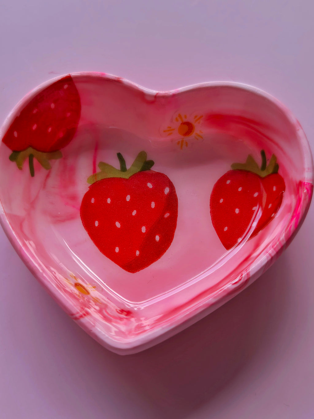 A heart-shaped pink trinket dish decorated with red strawberries and a small yellow flower, shown on a light pink background.