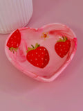 A heart-shaped pink trinket dish with decoupage strawberries and a small yellow flower, shown on a pink surface with part of a white ribbed container visible in the upper left.