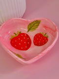 A heart-shaped pink trinket dish decorated with two red strawberries and green leaves, set against a soft pink background.