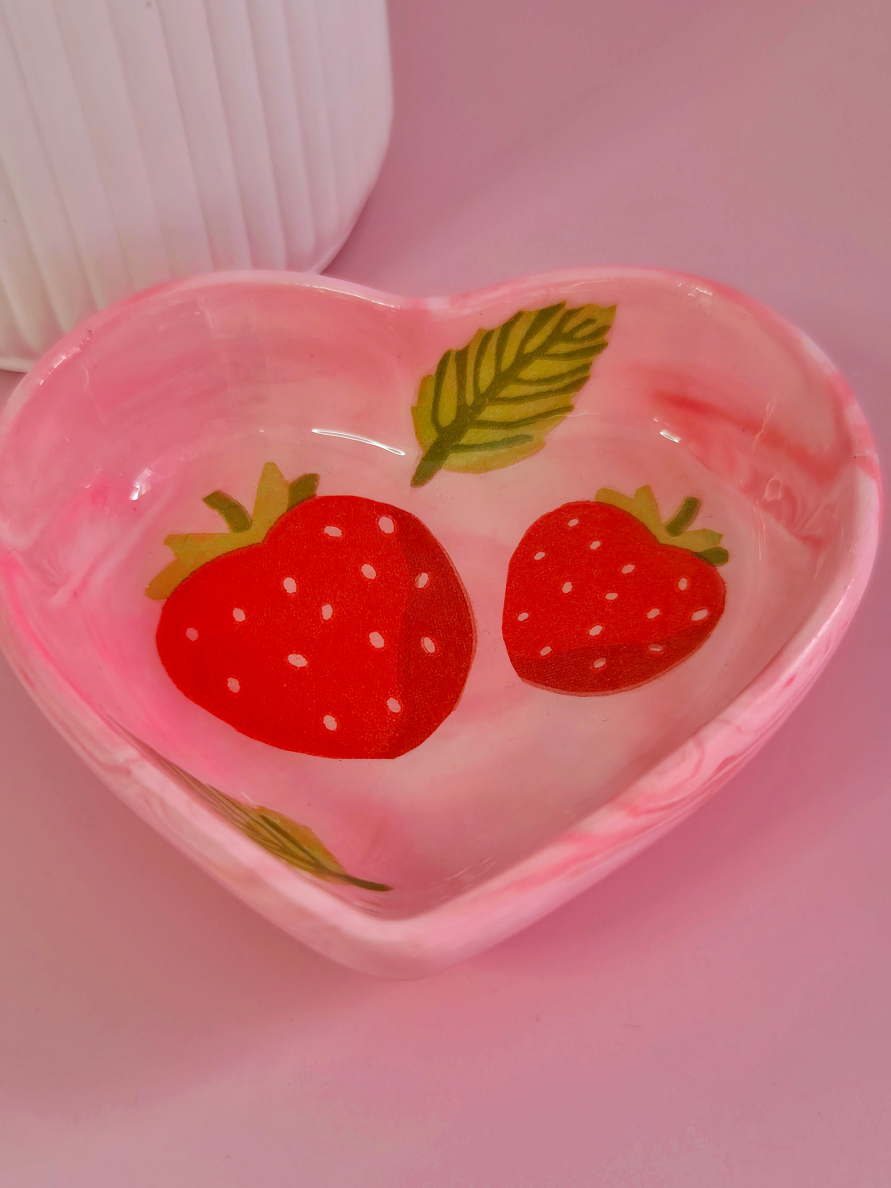 A heart-shaped pink trinket dish decorated with two red strawberries and green leaves, set against a soft pink background.