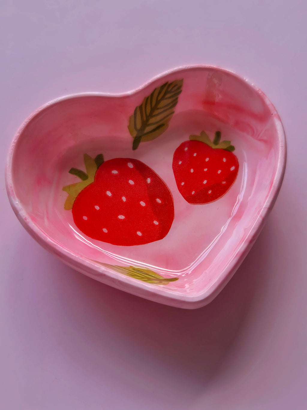 Handmade pink strawberry heart trinket dish shown at an angle, featuring a glossy sealed finish, red strawberries and soft marbled pink tones.