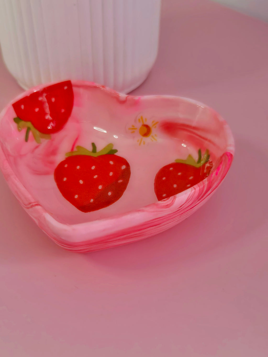 A small heart-shaped pink trinket dish with a swirled marbled pattern and decoupage red strawberries, shown on a pink surface with a white ribbed vase in the background.