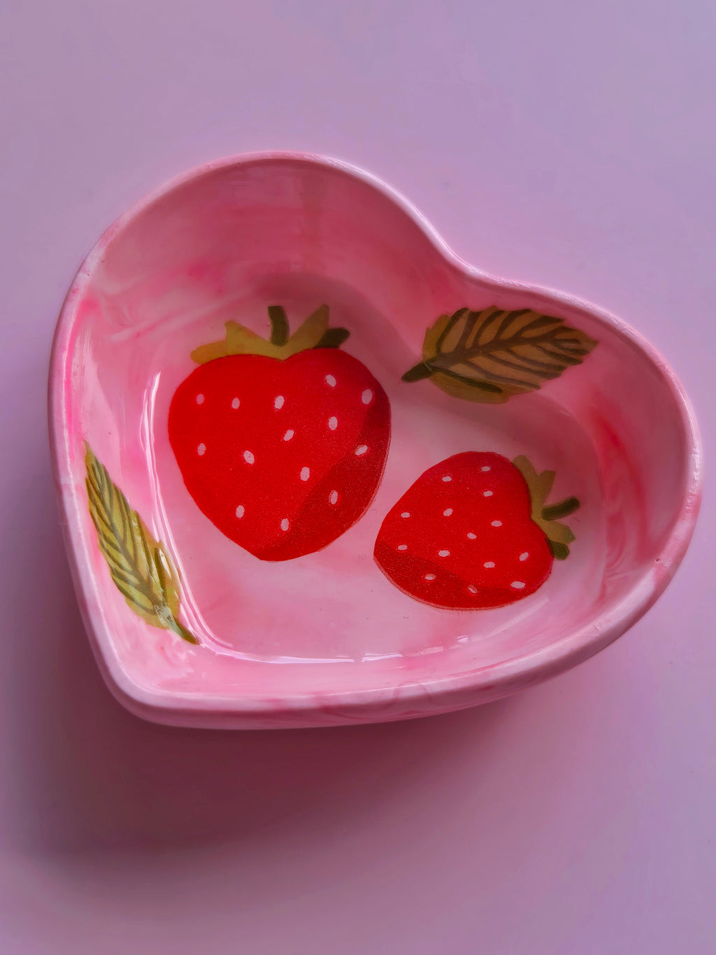 Top view of a pink heart-shaped trinket dish with two red strawberries, a green leaf design and a smooth glossy resincrete finish.