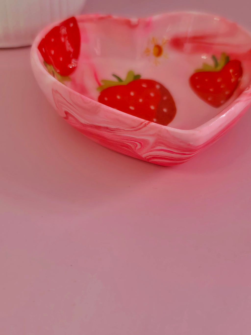 A small heart-shaped trinket dish with a pink and white marbled pattern, decorated with red strawberries and green leaves, set against a solid pink background.