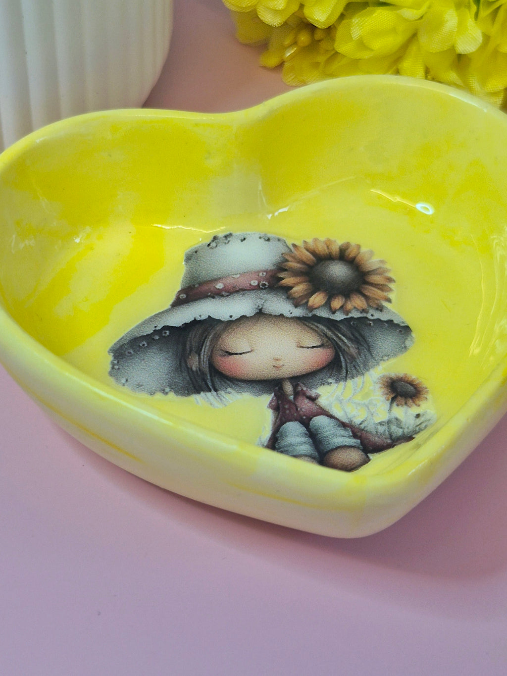 Heart-shaped yellow trinket dish with a girl and sunflower design on a pink surface side view 