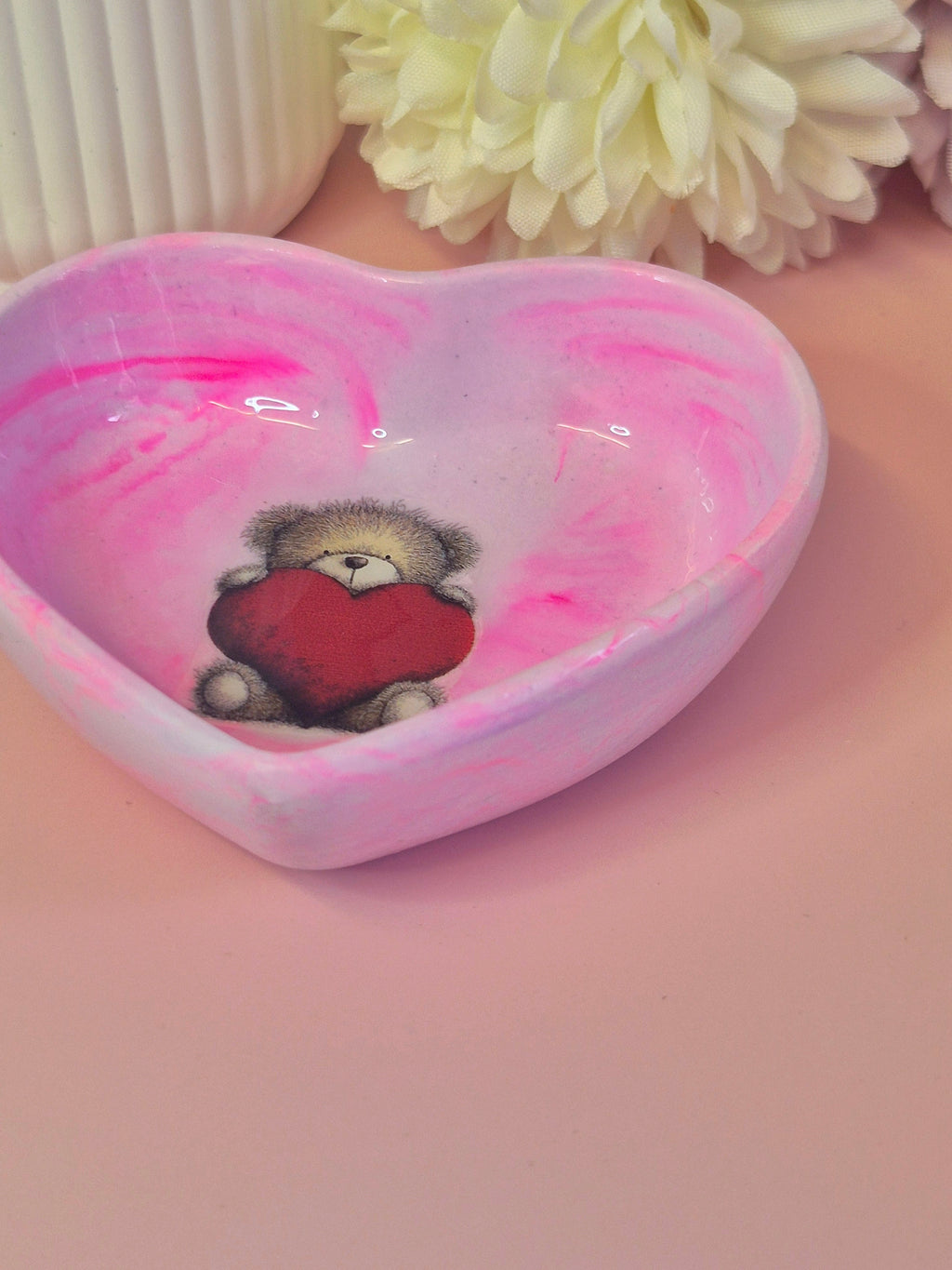 Heart-shaped trinket dish with a teddy bear holding a heart on a pink surface with flowers in the background