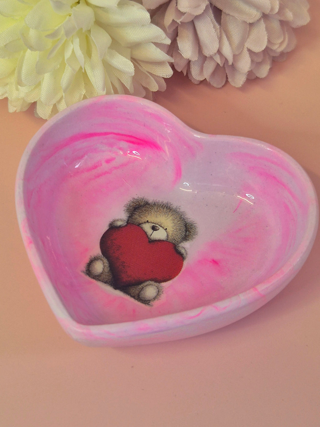 Heart-shaped pink trinket dish with a teddy bear holding a red heart, surrounded by light-colored flowers.